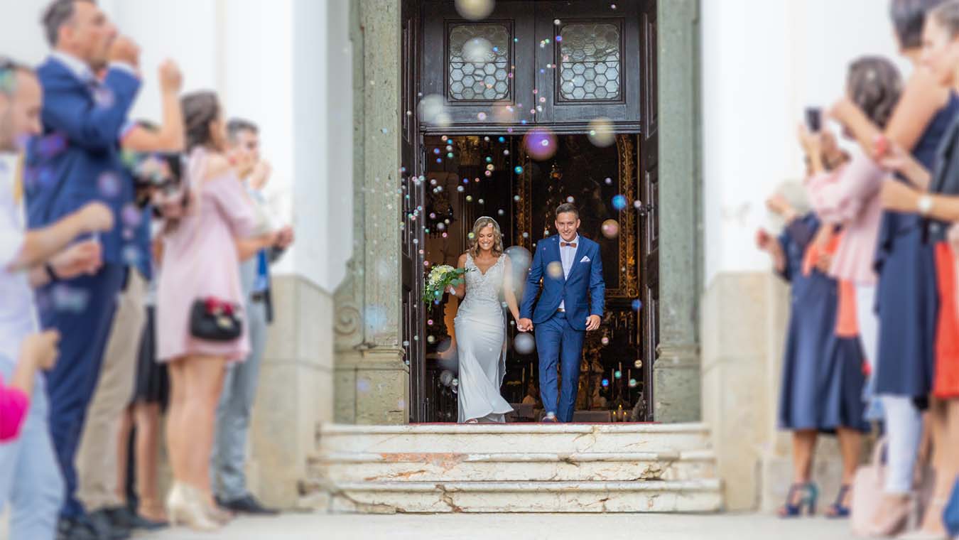 Bride and groom walk down church steps after their wedding while guests blow bubbles.
